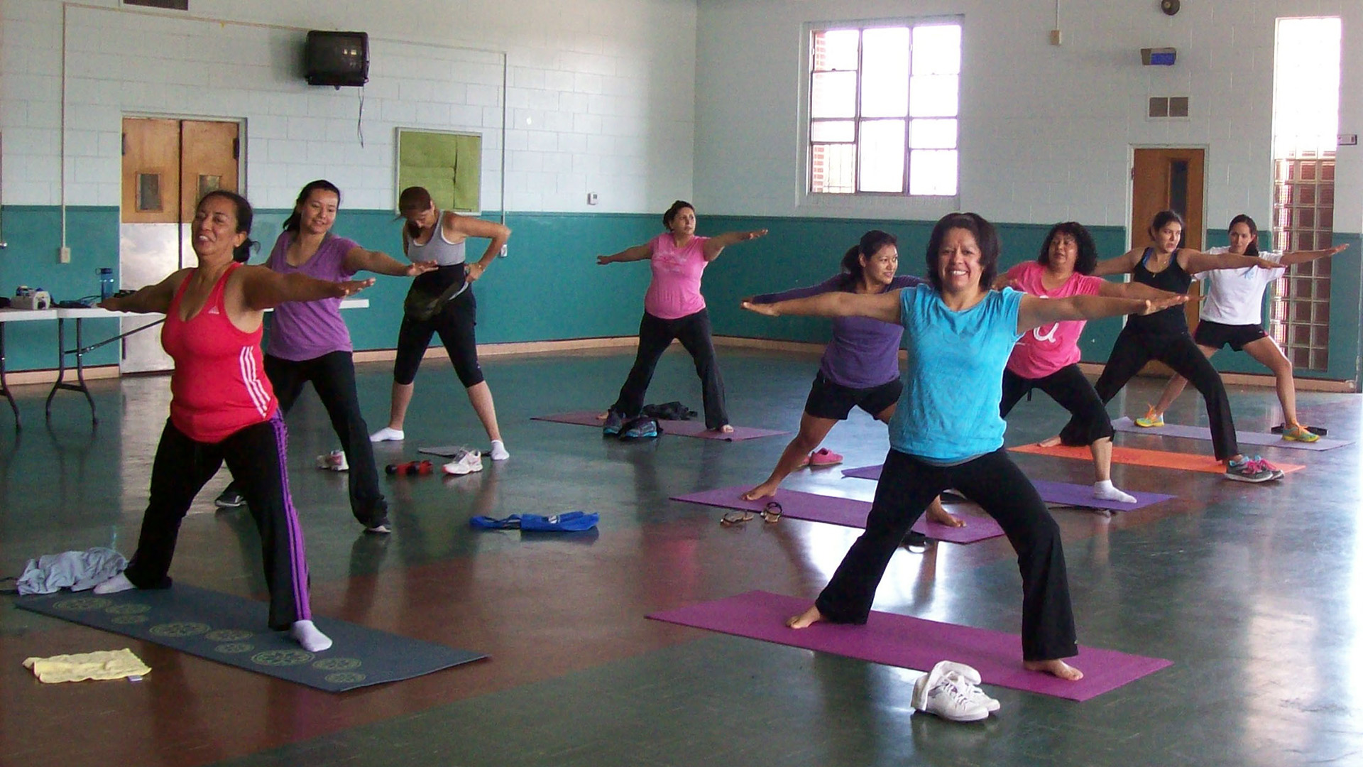 Nine Latinas in colorful workout attire standing on mats with their arms out to their sides.