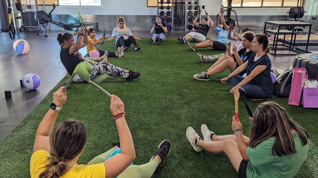 Latina mothers and children sit in a circle in YMCA gym, playing active games together.